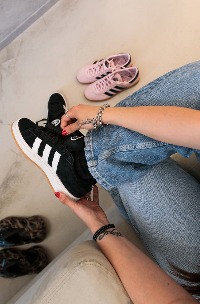 Person wearing black Adidas sneakers while sitting on a couch, with pink shoes visible in the background.