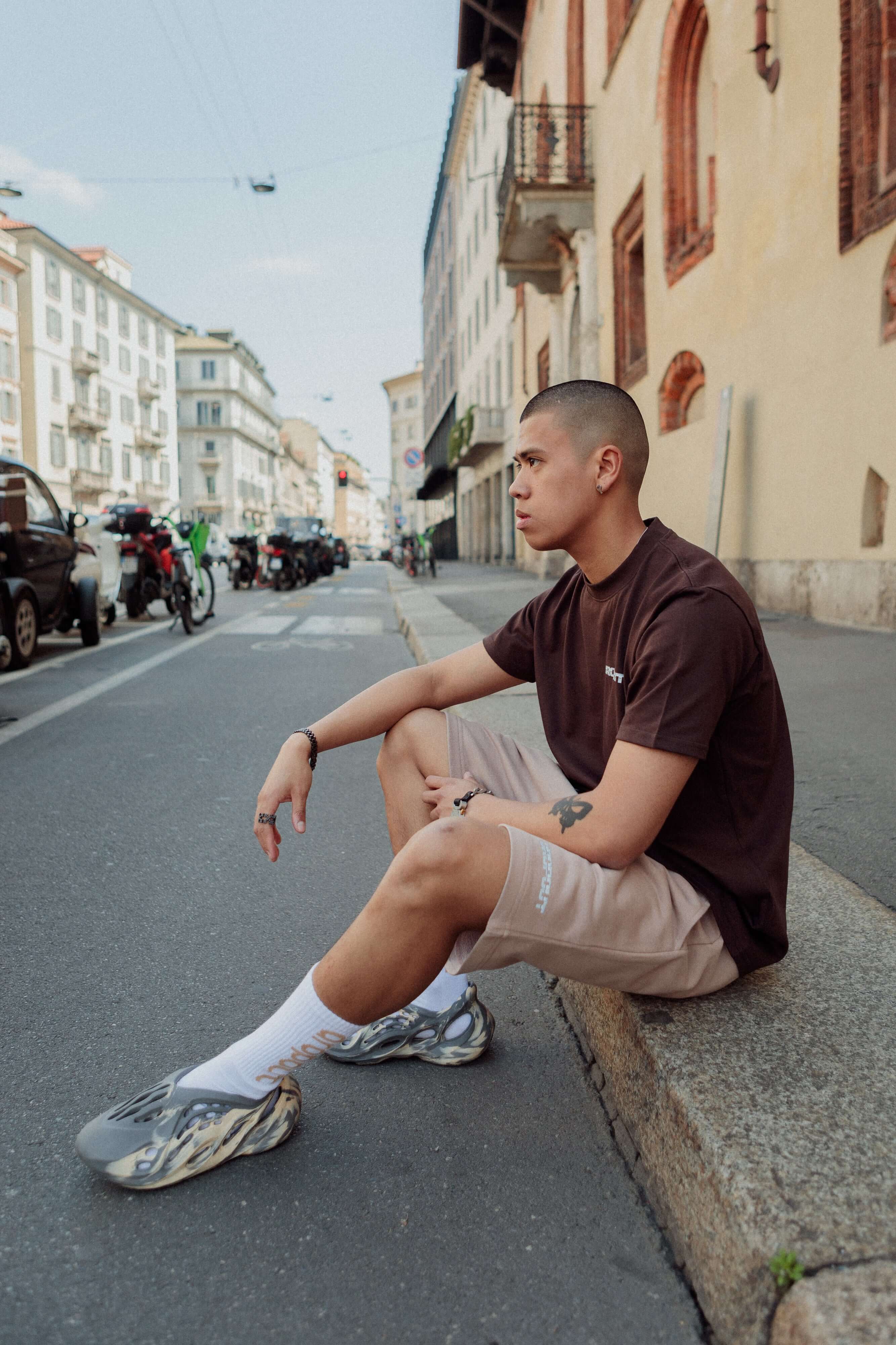 Young man sitting on a city street, wearing a brown t-shirt and beige shorts, with stylish sneakers.