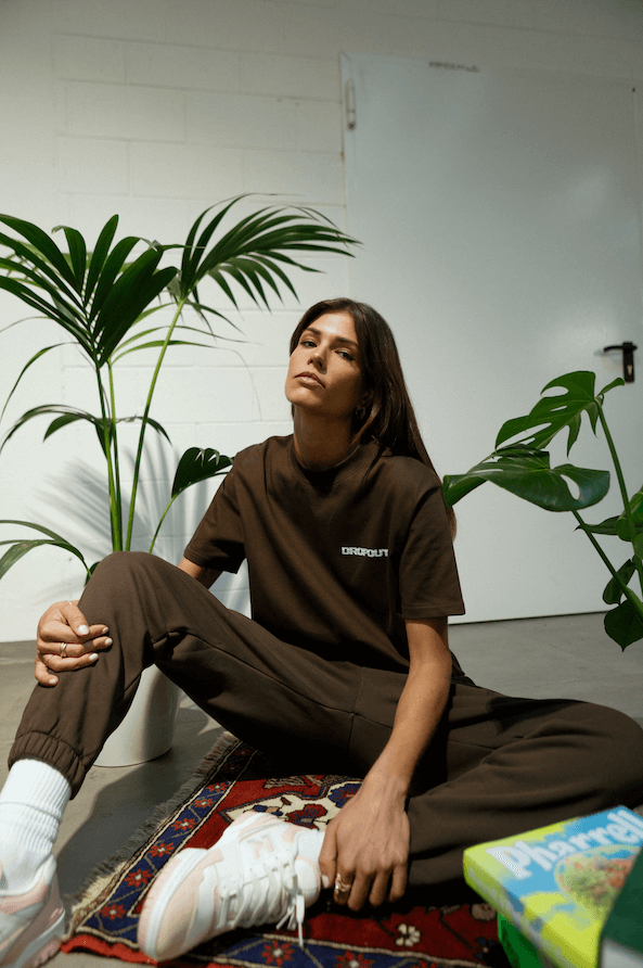 Fashionable model in brown outfit sitting with plants in a stylish interior, showcasing a relaxed look.