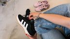 Person lacing up adidas Campus 00s Core Black sneakers while pink pair is in the background on a light surface.