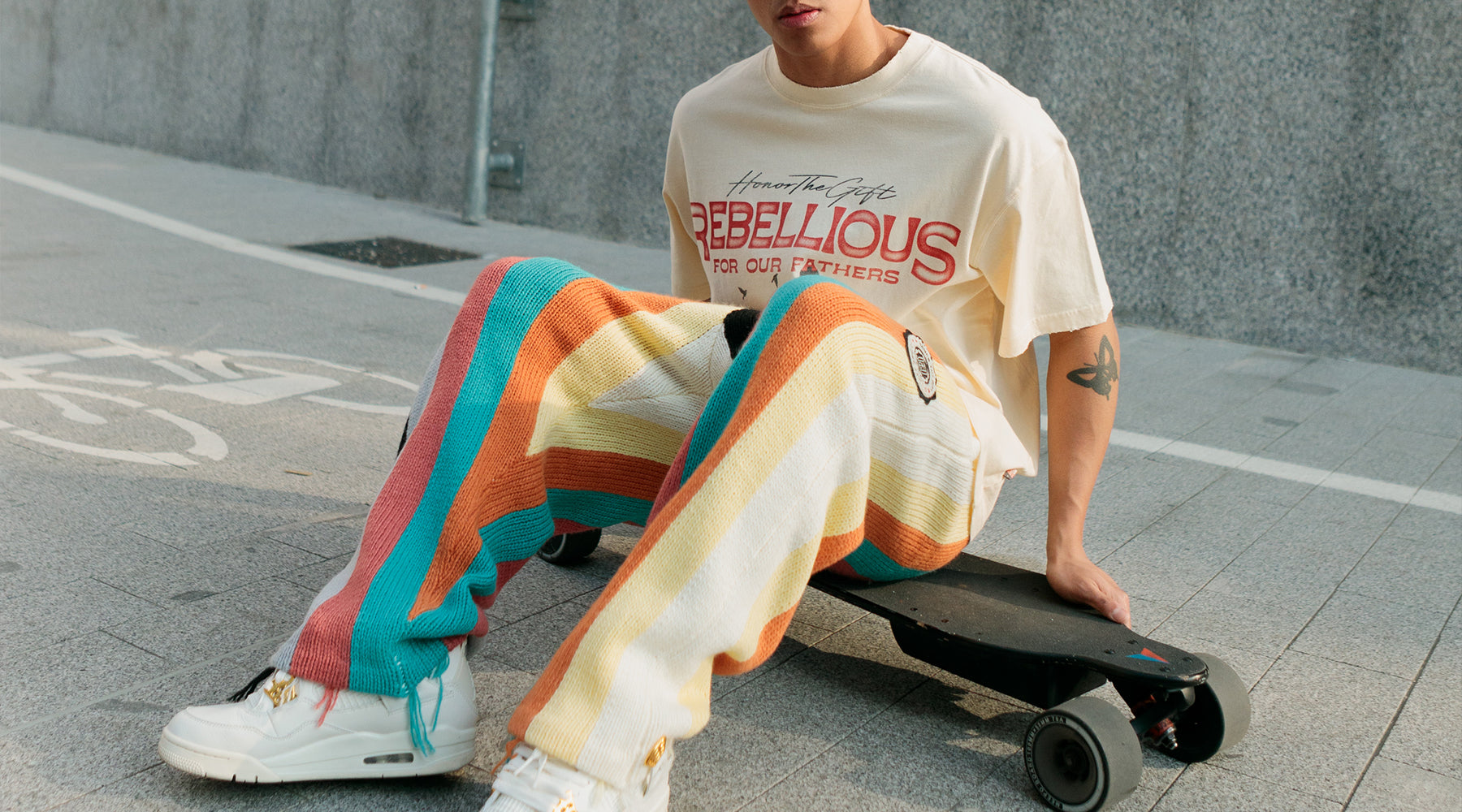 Person sitting on a skateboard, wearing a cream rebellious graphic t-shirt and colorful striped pants.